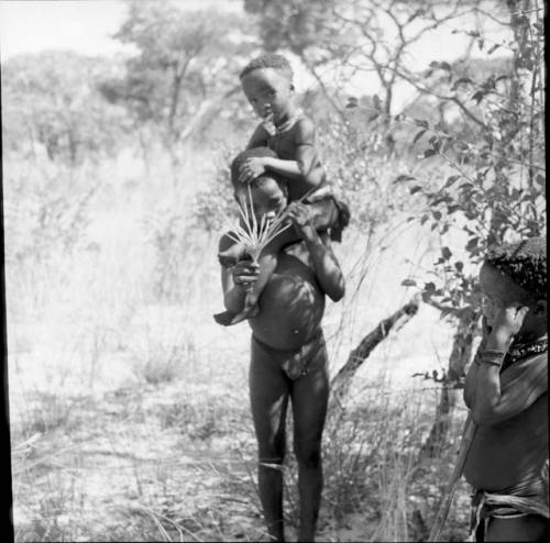 Boy holding !Ungka Norna on his shoulders, holding a palmate plant in his hand, with another child standing near them