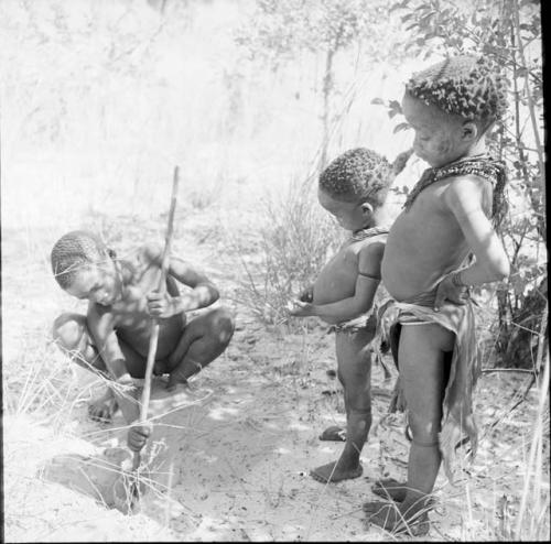 Boy digging in the sand with his digging stick, with !Ungka Norna and another child watching him