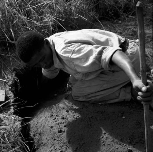 "Short /Qui" kneeling, reaching into a hole, holding his digging stick