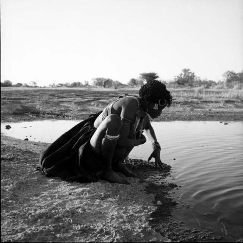 Woman wearing a kaross around her waist, squatting by a pan, looking in the water