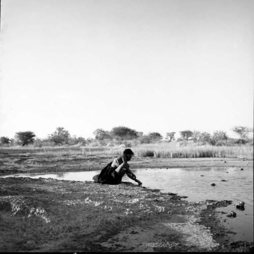 Woman wearing a kaross around her waist, squatting by a pan, with her hand in the water