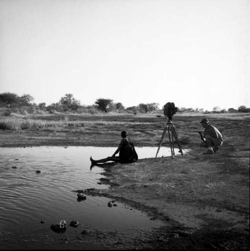 Woman sitting at the edge of a pan, bathing, with Nicholas England squatting next to a camera on a tripod near her