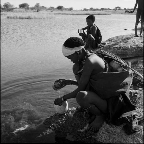 N!ai wearing a kaross, squatting at the edge of a pan, pouring water from a tin into an ostrich eggshell, with a bag of ostrich eggshells on her back, a person squatting behind her