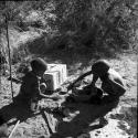 Two children play with a toy car made from a file box, with a carton and tin on the ground next to them