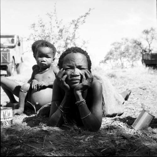 Woman lying on the ground, with her head in her hands, a child standing next to her, expedition Jeep and truck in the background