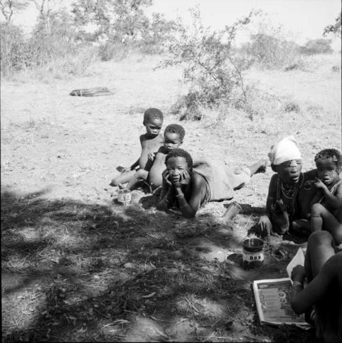 Woman lying on the ground, with her head in her hands, a child standing next to her, woman and children sitting near her, magazine lying on the ground next to them