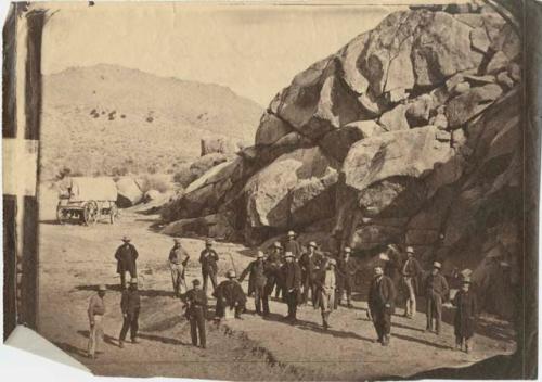 Group of men in front of a rock outcrop, with covered wagon in background