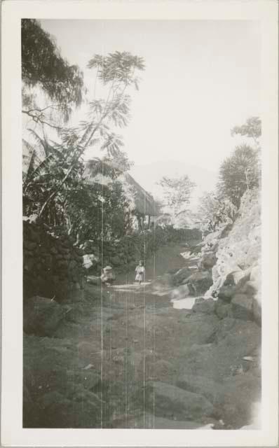 Child on rock wall path, view of thatched-roof building behind trees