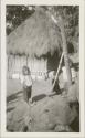 Three children standing in yard, thatched-roof behind them