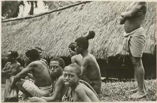Group of men and boys sitting in front of a thatched building