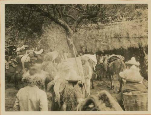 People and animals grouped near a thatched-roof building