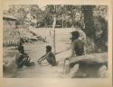 Three men sitting, with thatched structure in background