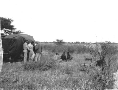 People sitting in the grass, being photographed by an expedition member, with other expedition members standing next to the trucks near them