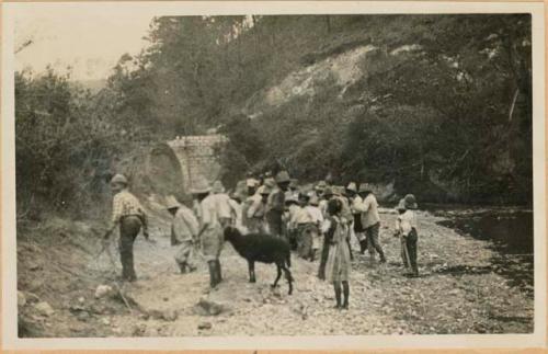 Group on bank of river, with stone bridge in background