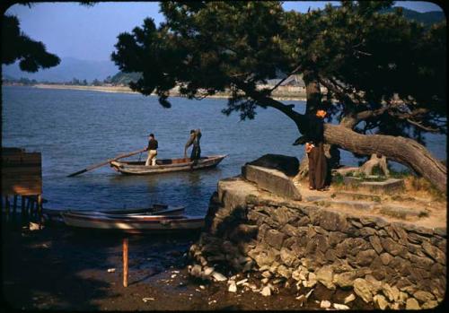 Two men in fishing boat on river, and man standing under tree on shore