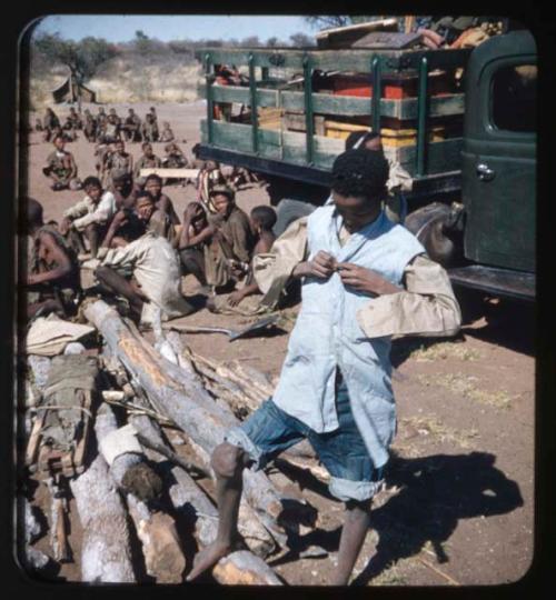 Expedition: Boy putting on Western clothing, with a group of people sitting by a truck and pile of wood in the background