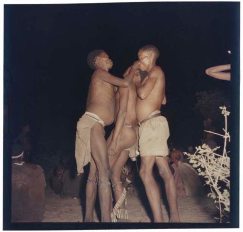 Three men standing, one in a trance, at a night dance