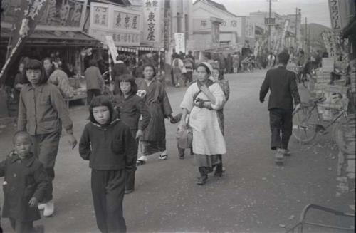 Group of women and children walking down street, with storefronts in background