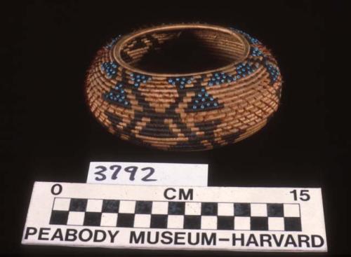 Small coiled basket, woven design in black, feather and bead ornament.