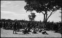 Group of men sitting and standing under a tree and by a fence