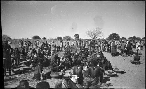 Group of people sitting and standing, with baskets of grain on the ground near them