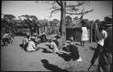 Men sitting near a tree, and a woman carrying a pot on her head