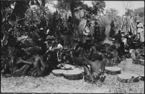 Group of people standing and sitting under trees, with a large basket on its side and shallow baskets