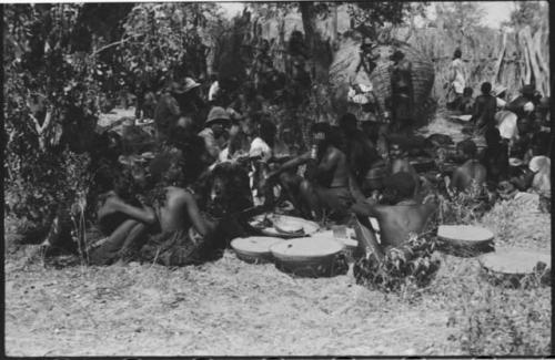 Group of people standing and sitting under trees, with a large basket on its side and shallow baskets