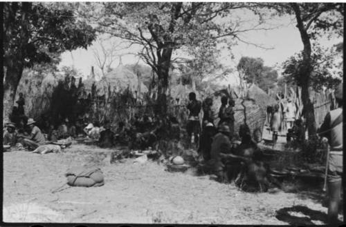 Group of people standing and sitting under trees, with a large basket on its side