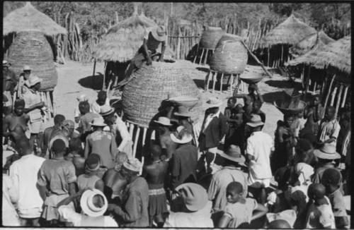 Group of people standing in front of storage baskets, and a man sitting on top of one