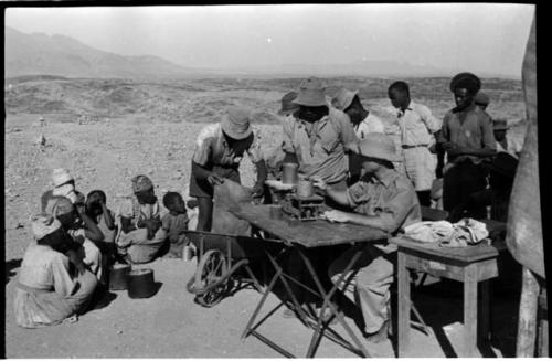 Group of people sitting and standing around a man sitting at a table weighing tin ore