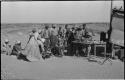 Group of people sitting and standing around a man sitting at a table weighing tin ore