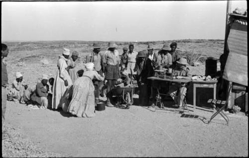 Group of people sitting and standing around a man sitting at a table weighing tin ore