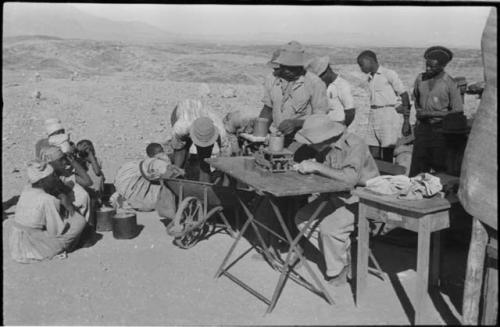 Group of people sitting and standing around a man sitting at a table weighing tin ore