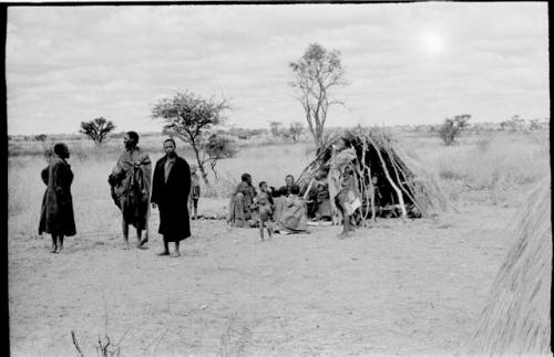 Three men standing (two wearing long coats), next to a group sitting in front of a hut; woman standing wearing a short kaross