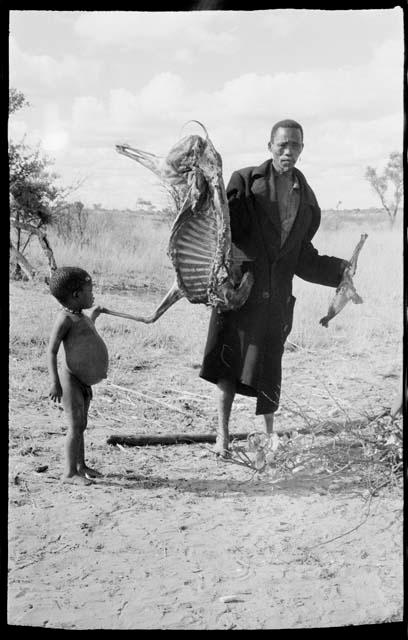 Man in a black overcoat holding up half an animal carcass (which the Marshalls gifted); a child standing next to him