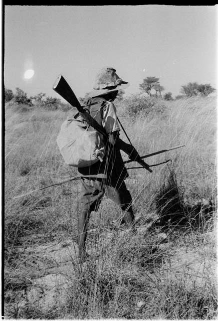 Headman walking, carrying a gun, a spear and a bundle, view from behind