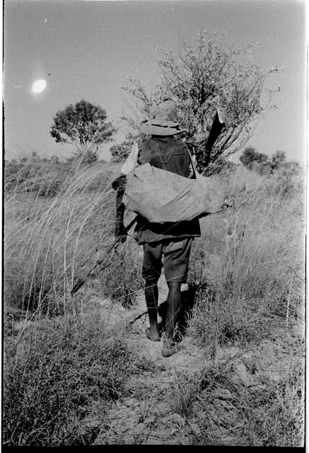 Headman walking, carrying a gun, a spear and a bundle, view from behind