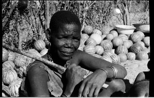 Woman sitting holding a long stick in her hands; tsama melons in the background