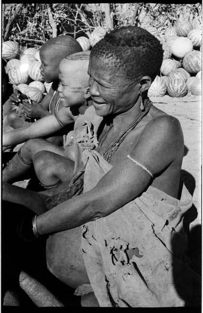 Elderly woman and two children sitting in a row; tsama melons in the background