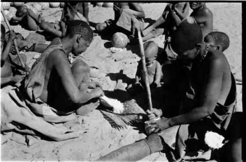 Woman sieving millet using a mat; woman (who is Kgalagari) opening a tsama melon with a digging stick; children and people in the background