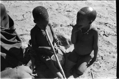 Two children sitting with a tsama melon; one child sucking end of a digging stick