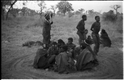 Three men, including /Gunda and ≠Gao, dancing around a group of seated women