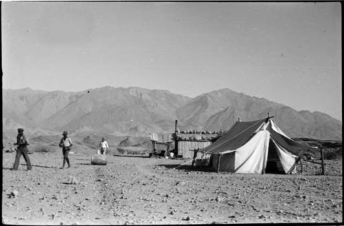 Men walking through a camp with a tent and corrugated metal shed