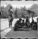 Girls sitting and working on a woven band with beads on it, and a woman standing next to them