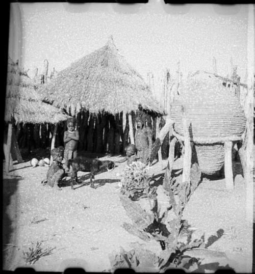 Children sitting and standing with a goat near storage huts inside kraal