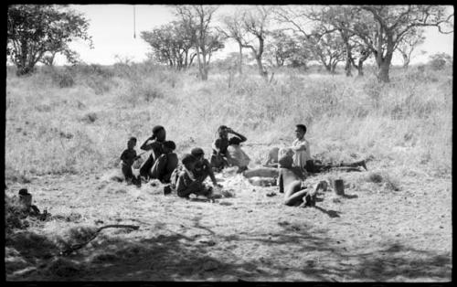 A group of people seen from a distance, sitting in the sun, including !U with !Ungka Norna in her lap, Di!ai, and her son /Gaishay beside her, Debe standing beside !Ungka Norna, an unidentified, visiting girl, !Ungka, and "≠Gao Lame"