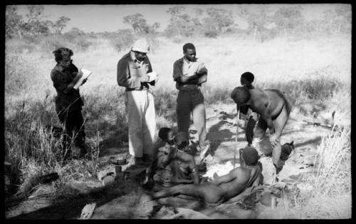 "Old Gau" standing with his bandaged foot in a bucket, with Elizabeth Marshall Thomas, Robert Story, Kernel Ledimo, and other unidentified people present