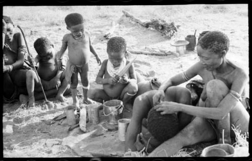 Unidentified person and Bau, who is playing with a digging stick to the left, Debe watching /Gaishay cut a piece of meat with a knife, and /Naoka looking through "Gao Medicine's" hair for lice; part of Elizabeth Marshall Thomas's child study