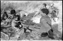 !Ungka Norna holding a piece of meat, seated between !U and ≠Toma with two unidentified people in the background